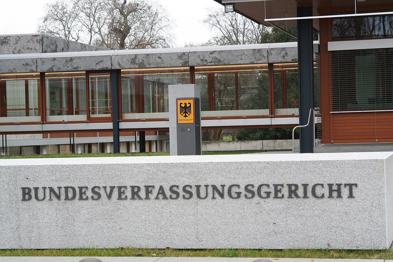Foto: Eine helle Steinmauer mit dem Schriftzug Bundesverfassungsgericht, dahinter ein flaches Gebäude mit viel Glas und eine Stele mit Bundesadler.