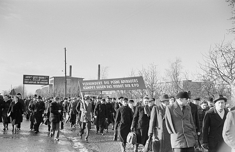 Schwarz-Weiß-Foto von 1952: Ein langer Zug von Arbeitern in Mänteln läuft über eine Straße. Auf einem Transparent steht: Verhindert die Pläne Adenauers, kämpft gegen das Verbot der KPD.