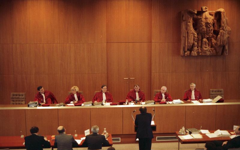 Foto: Richterinnen und Richter in roten Roben sitzen an der Richterbank, hinter ihnen eine holzgetäfelte Wand mit Bundesadler. Vorn steht ein Mann am Rednerpult.